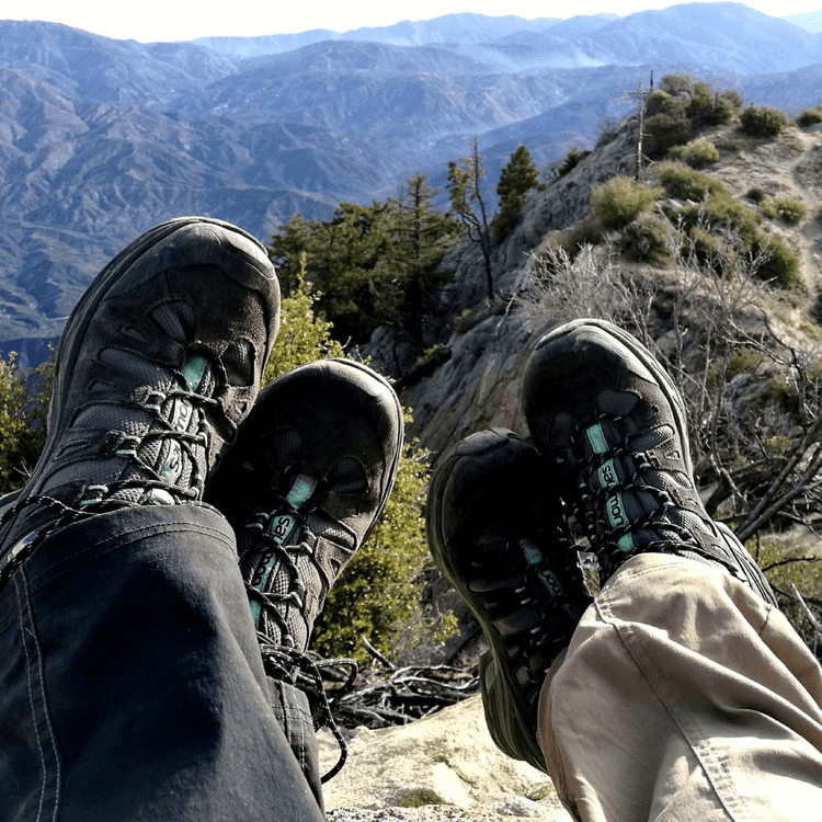 Two pairs of hiking boots resting on a rocky ledge with mountains in the background, showcasing outdoor adventure footwear Collection, Adventure Travel Gear