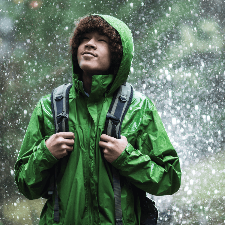 Person wearing a green waterproof jacket in the rain, smiling with a backpack, Jackets Collection Adventure Travel Gear